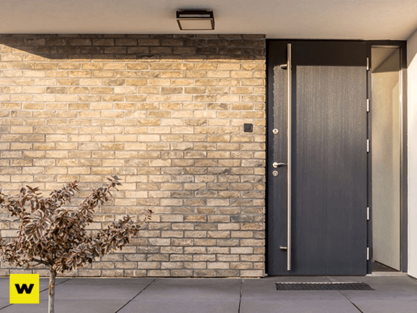 Moderne Haustür aus dunklem Holz neben heller Klinkerfassade mit kleinem Strauch davor.