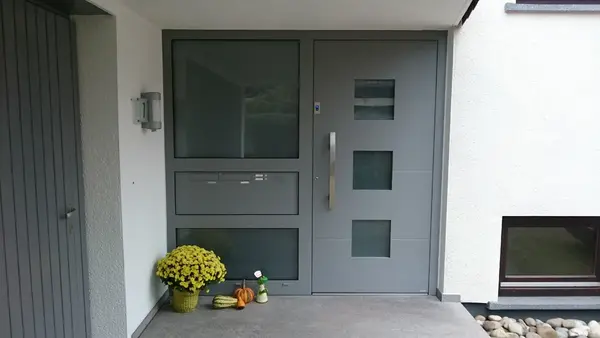 Moderne graue Haustür mit seitlichem Glaselement, gelben Blumen und Kürbissen im Eingangsbereich.